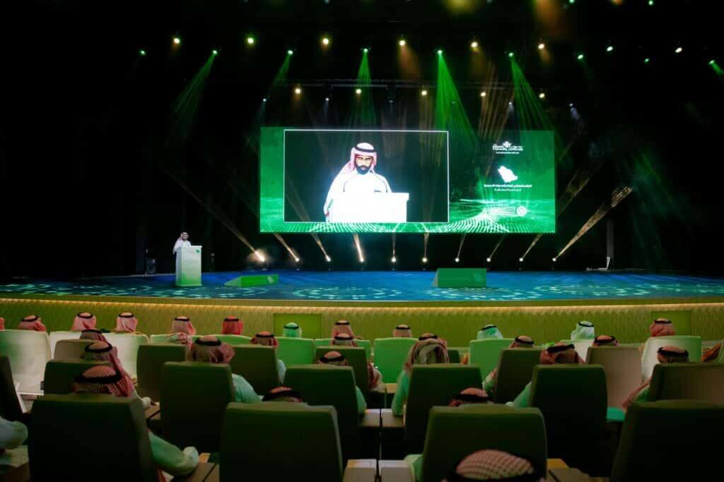 Delivering a speech at the GeoGeography Conference at Qassim University with an academic audience in a green-lit hall.