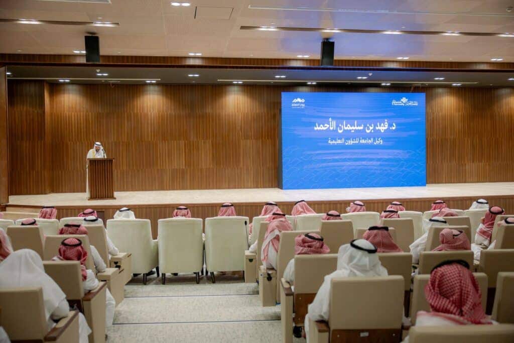 إلقاء كلمة في قاعة جامعة القصيم خلال فعالية اليوم العالمي للمعلم، مع شاشة شاشة توضح المناسبة.