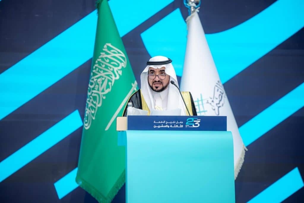 Delivering a formal speech at the graduation ceremony of the 23rd class of Qassim University in front of a blue background.
