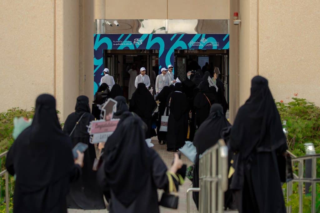 An academic scene in front of the entrance to Qassim University suggests a graduation ceremony and an active student presence.
