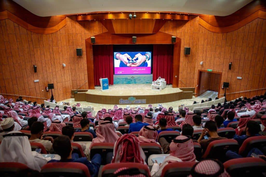 Academic conference at Qassim University with a large attendance, featuring speakers and presentations on stage.