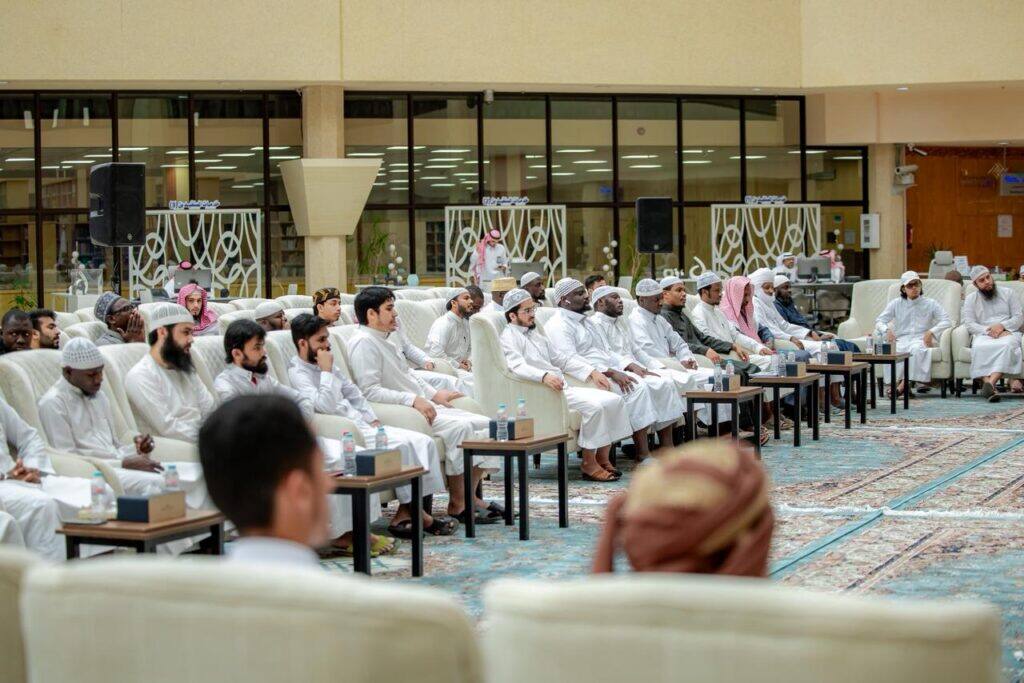 An academic meeting for students of Qassim University in a hall with tables in the center and the library in the background.